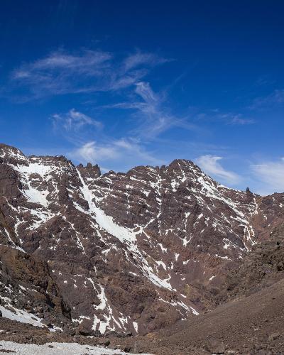 A high-altitude mountain valley with steep rocky slopes and patches of snow under a blue sky.