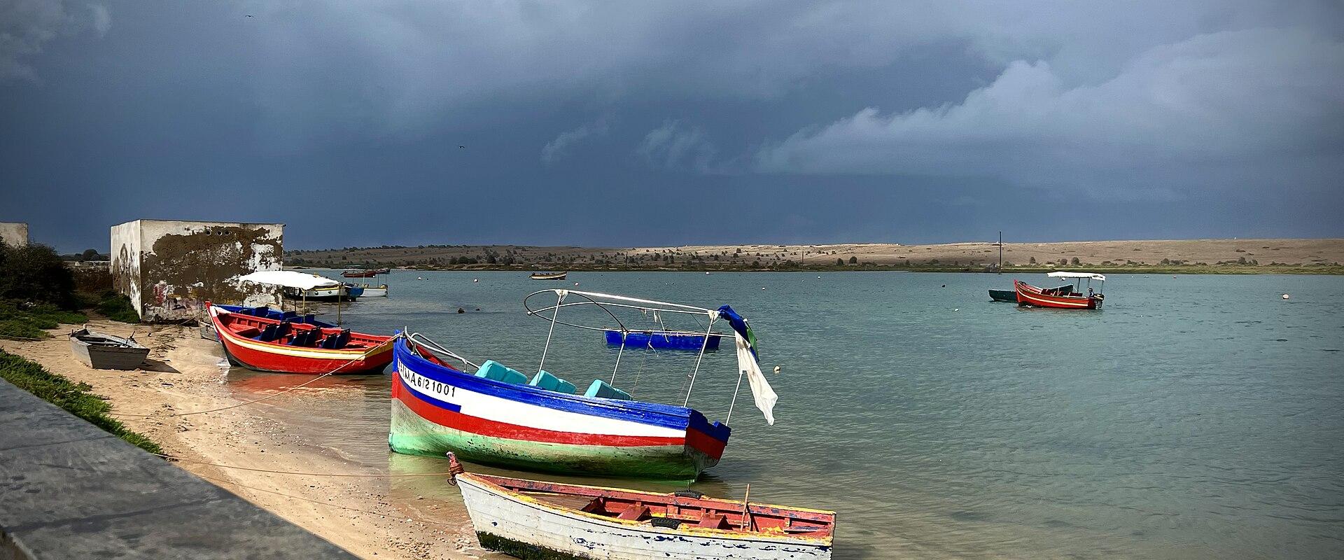 A coastal scene showing several colorful fishing boats moored in a calm lagoon or estuary under a dramatic, cloudy sky, with a sandy shore and a weathered building on the left.