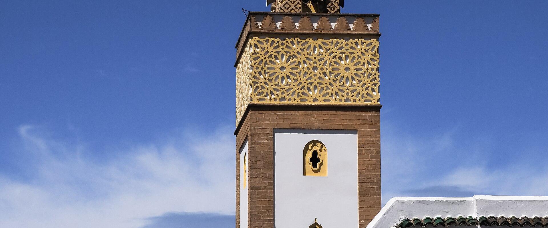 A close-up view of a mosque minaret featuring intricate golden geometric latticework, brickwork, and a green-tiled roof against a blue sky.