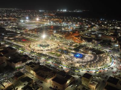 Aerial night view of a brightly lit urban plaza featuring a large central fountain and monument, surrounded by city streets and buildings, with an airport runway visible in the background.