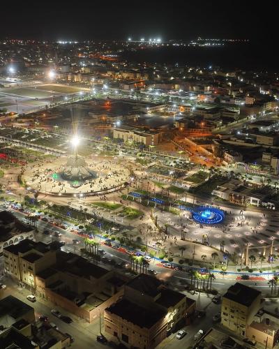 Aerial night view of a brightly lit urban plaza featuring a large central fountain and monument, surrounded by city streets and buildings, with an airport runway visible in the background.