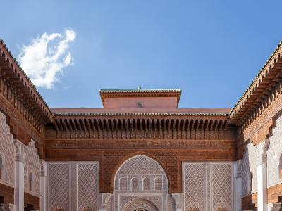 A low-angle photograph of an ornate courtyard featuring intricate stucco carvings, carved cedar wood eaves, and a central horseshoe archway under a blue sky.