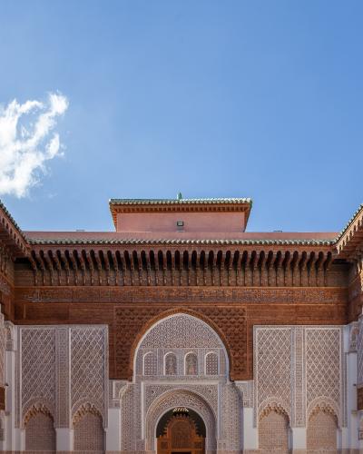 A low-angle photograph of an ornate courtyard featuring intricate stucco carvings, carved cedar wood eaves, and a central horseshoe archway under a blue sky.