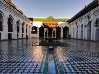 A courtyard with white walls, horseshoe arches, a central fountain, and a distinctive green-tiled roof structure in the background.