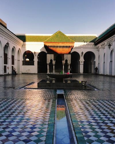A courtyard with white walls, horseshoe arches, a central fountain, and a distinctive green-tiled roof structure in the background.