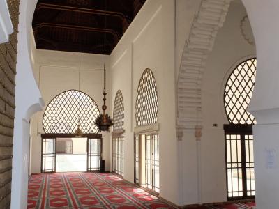 The interior of a mosque or prayer hall featuring red patterned carpets, white walls, horseshoe arches, and a dark wooden ceiling with hanging lanterns.