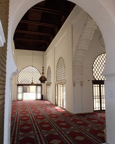 The interior of a mosque or prayer hall featuring red patterned carpets, white walls, horseshoe arches, and a dark wooden ceiling with hanging lanterns.