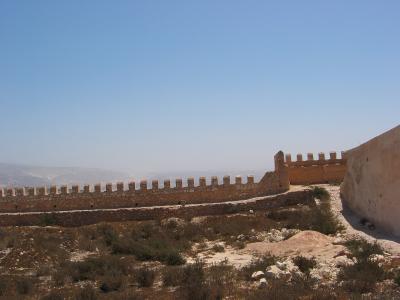 The image shows the stone ramparts and crenellated walls of a historic fortress situated on a dry, scrub-covered hilltop under a clear blue sky.