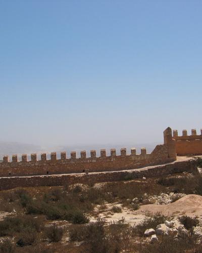 The image shows the stone ramparts and crenellated walls of a historic fortress situated on a dry, scrub-covered hilltop under a clear blue sky.