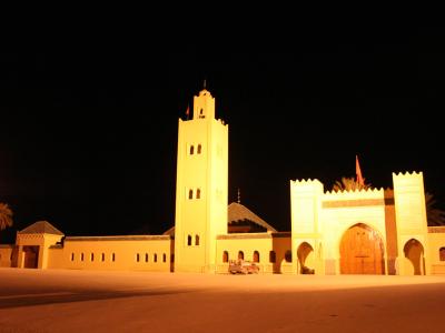 A night photograph of a large, illuminated religious complex featuring a tall square minaret, a domed structure, and a fortified entrance gate with a Moroccan flag, set against a dark sky with palm trees visible.