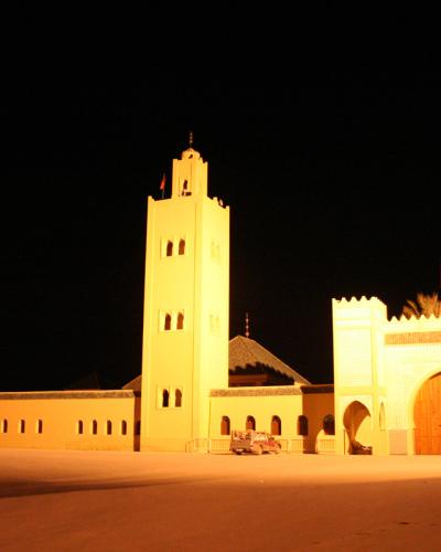 A night photograph of a large, illuminated religious complex featuring a tall square minaret, a domed structure, and a fortified entrance gate with a Moroccan flag, set against a dark sky with palm trees visible.