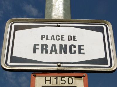 A close-up of a white rectangular street sign with black text reading "PLACE DE FRANCE" mounted on a metal pole against a blue sky, with a smaller orange sign below it labeled "H150".
