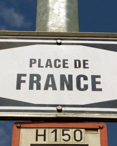 A close-up of a white rectangular street sign with black text reading "PLACE DE FRANCE" mounted on a metal pole against a blue sky, with a smaller orange sign below it labeled "H150".