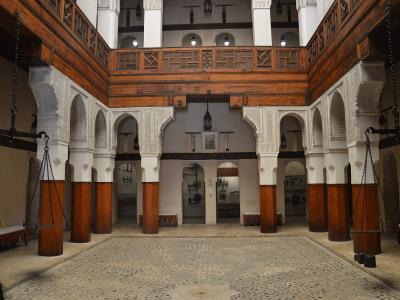 The image shows the interior courtyard of a traditional Moroccan funduq (caravanserai) with ornate wooden balconies, carved stucco arches, and large hanging scales, displaying wooden artifacts in the background.