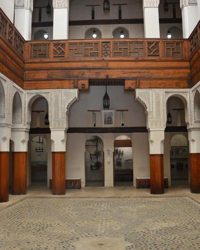 The image shows the interior courtyard of a traditional Moroccan funduq (caravanserai) with ornate wooden balconies, carved stucco arches, and large hanging scales, displaying wooden artifacts in the background.