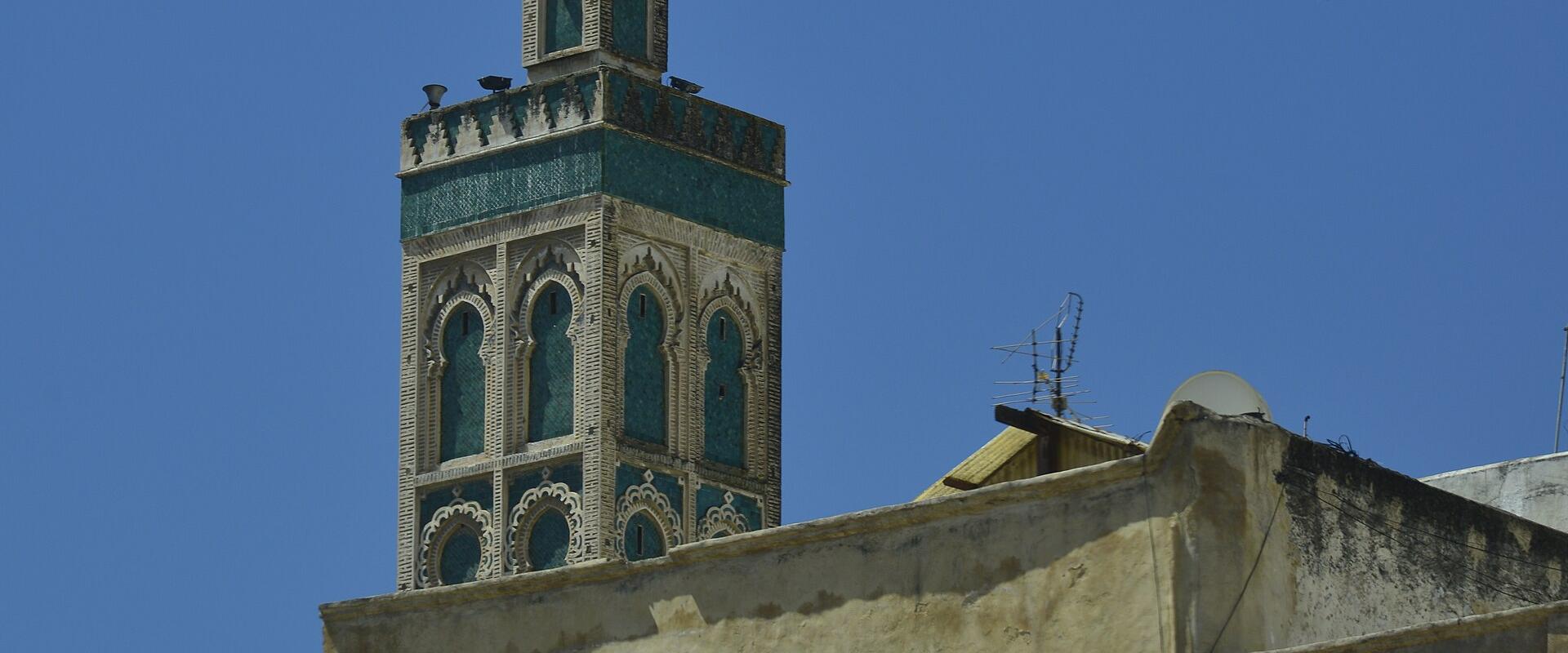 A tall, ornate minaret with green and white tilework rises above a beige building under a clear blue sky; the foreground shows part of a white vehicle with text reading "NAVETTE SPECIALE RCIF" in French and Arabic.