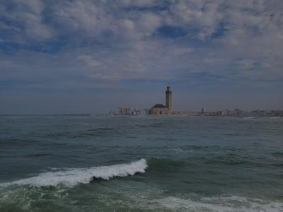 A distant view of a large mosque with a prominent minaret situated on a promontory extending into the ocean, with a city skyline in the background and a rocky shoreline in the foreground.
