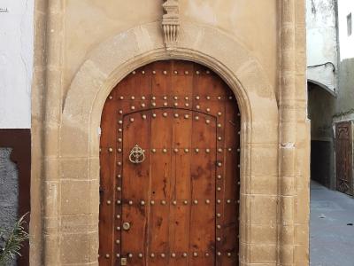 A close-up photograph of a traditional arched wooden door with metal studs and a knocker, framed by a stone portal with columns and a keystone, set within a narrow street.