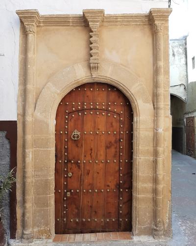 A close-up photograph of a traditional arched wooden door with metal studs and a knocker, framed by a stone portal with columns and a keystone, set within a narrow street.