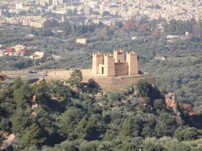 A reddish-brown fortress with four corner towers sits atop a green, tree-covered hill, overlooking a sprawling modern city in the background.