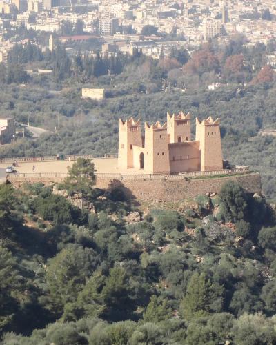 A reddish-brown fortress with four corner towers sits atop a green, tree-covered hill, overlooking a sprawling modern city in the background.