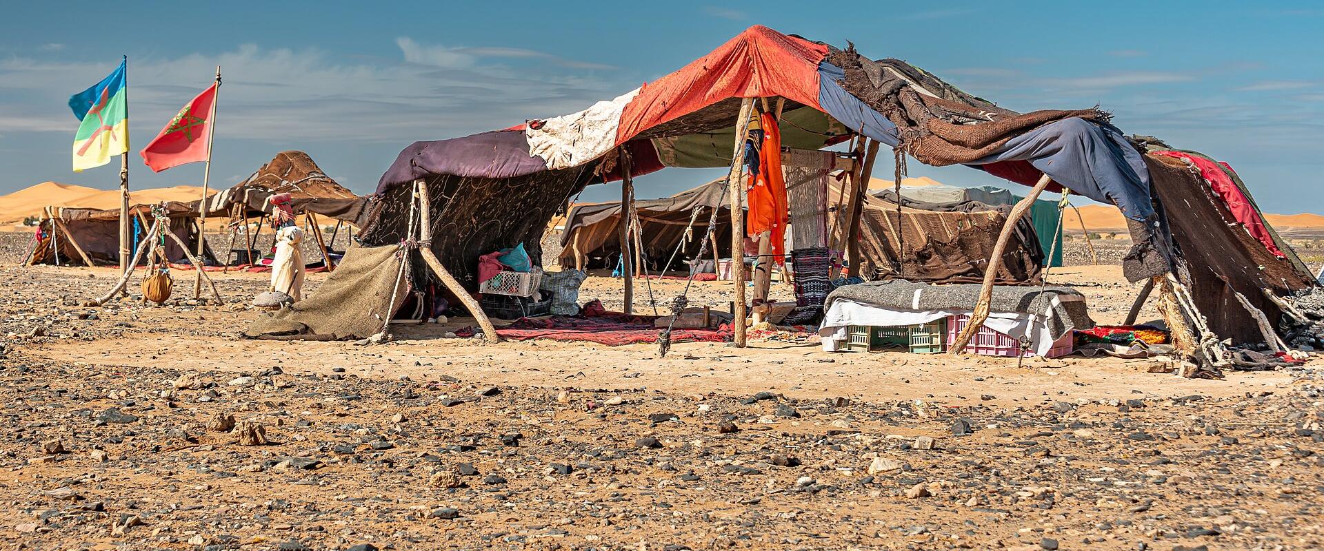 A makeshift desert encampment with tents constructed from fabric and wooden poles, set on rocky ground with sand dunes in the background, featuring a Moroccan flag and another flag on poles.