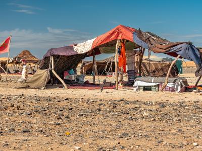A makeshift desert encampment with tents constructed from fabric and wooden poles, set on rocky ground with sand dunes in the background, featuring a Moroccan flag and another flag on poles.