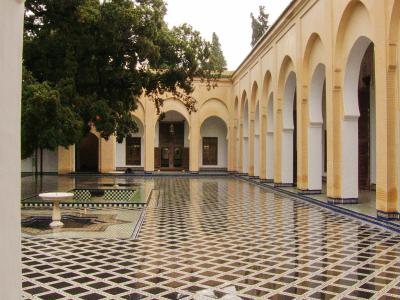 A courtyard with a large reflecting pool, black-and-white diamond-patterned tile flooring, and a colonnade of yellow walls with horseshoe arches.