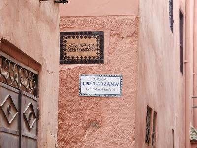 A narrow alleyway in a pink-walled medina showing two street signs: one reading "DERB FRANCISCO" and another reading "Synagogue 1492 'LAAZAMA' Derb Talmoud Thora 36".