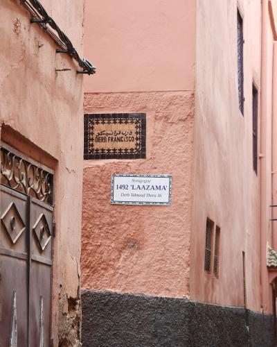 A narrow alleyway in a pink-walled medina showing two street signs: one reading "DERB FRANCISCO" and another reading "Synagogue 1492 'LAAZAMA' Derb Talmoud Thora 36".