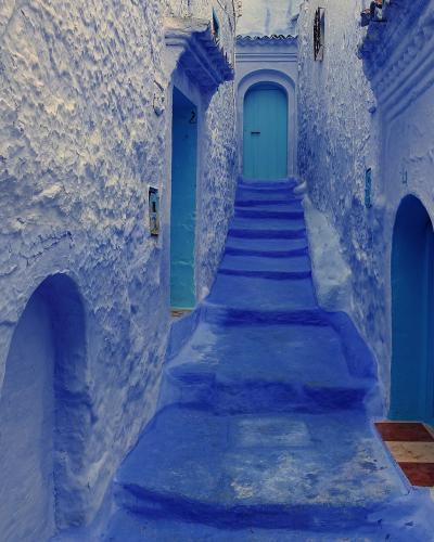 A narrow, steep alleyway within a medina, featuring textured white and blue-washed walls, arched doorways, and a central staircase leading to a blue door.