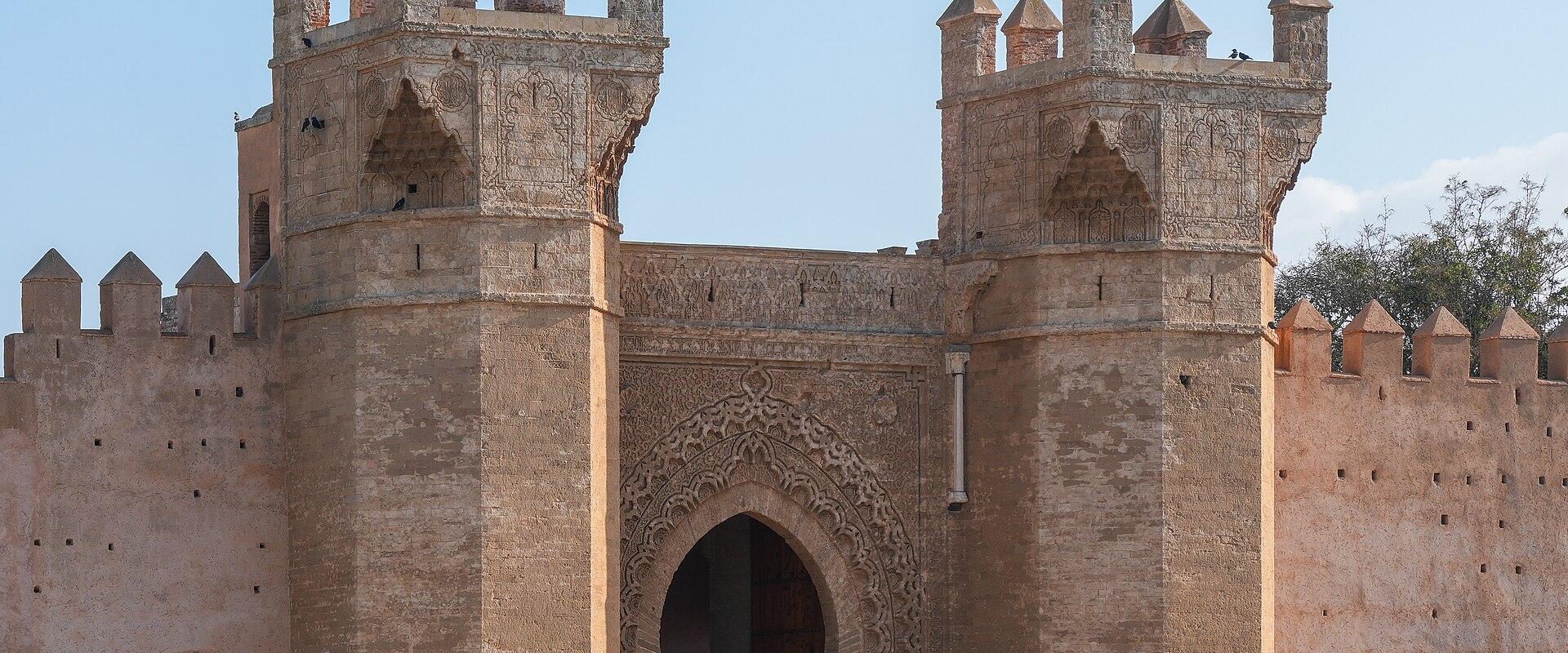 A large, ornate stone gateway with two prominent towers featuring crenellations and intricate stonework, set against a clear blue sky.