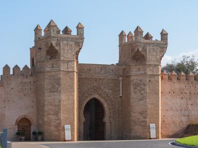 A large, ornate stone gateway with two prominent towers featuring crenellations and intricate stonework, set against a clear blue sky.