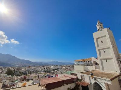 A high-angle view of a dense urban area with white buildings, a prominent white minaret with a distinctive crenellated top on the right, and mountains in the background under a bright blue sky.