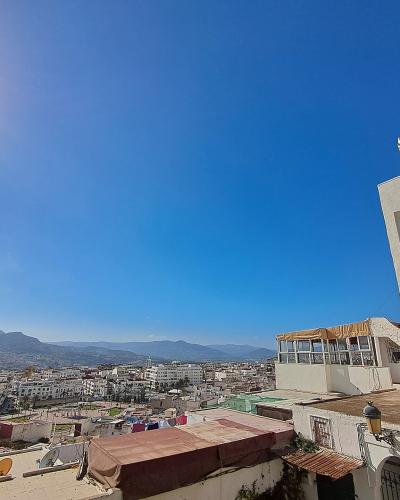 A high-angle view of a dense urban area with white buildings, a prominent white minaret with a distinctive crenellated top on the right, and mountains in the background under a bright blue sky.