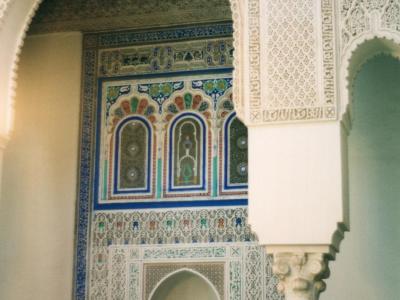 An interior architectural detail showing a horseshoe arch framing a wall decorated with intricate zellige tilework, carved stucco, and Arabic calligraphy, typical of Moroccan Islamic art.