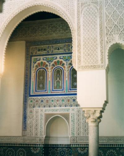An interior architectural detail showing a horseshoe arch framing a wall decorated with intricate zellige tilework, carved stucco, and Arabic calligraphy, typical of Moroccan Islamic art.