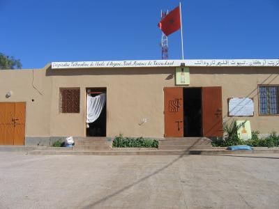 A single-story beige building with a Moroccan flag on the roof and a sign reading "Coopérative Taïmatine de l'huile d'Argane - Taroudant - Province de Taroudant" in French and Arabic.