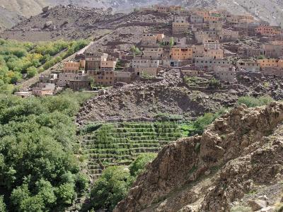 A village of terracotta-colored buildings built on a steep, rocky mountainside above green terraced fields and a stream, with rugged mountain peaks in the background.