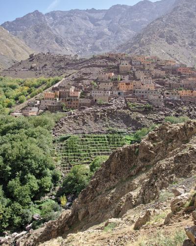 A village of terracotta-colored buildings built on a steep, rocky mountainside above green terraced fields and a stream, with rugged mountain peaks in the background.