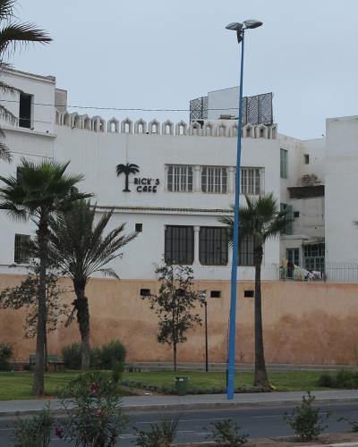 A white, multi-story building with a distinctive crenellated roofline and a palm tree logo on the facade, situated behind a tan wall and a row of palm trees along a street.