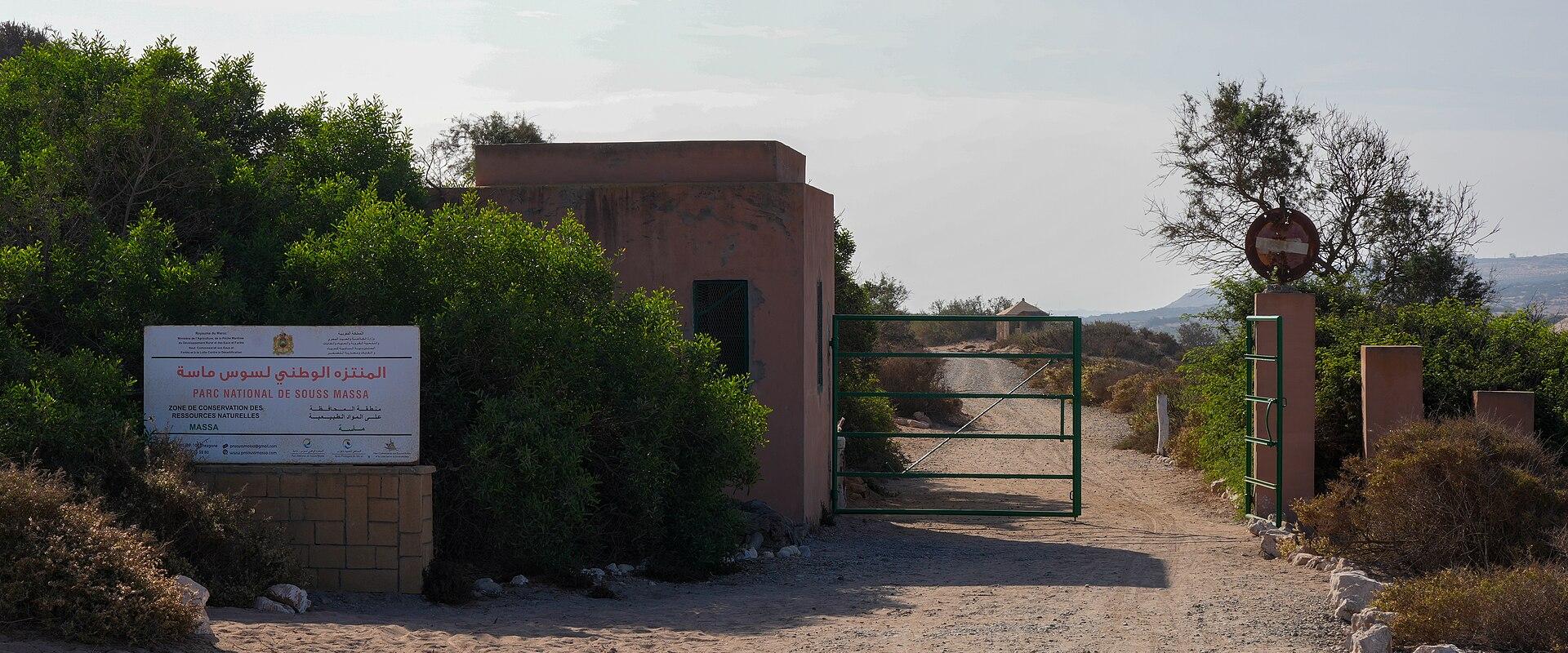 A dirt road entrance to a park featuring a guardhouse, a green metal gate, and a large white sign with Arabic and French text.