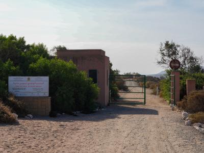 A dirt road entrance to a park featuring a guardhouse, a green metal gate, and a large white sign with Arabic and French text.