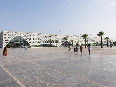 A wide shot of a modern train station building featuring a distinctive white geometric lattice facade, with people walking on a large paved plaza in front.