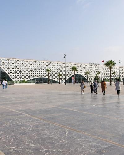 A wide shot of a modern train station building featuring a distinctive white geometric lattice facade, with people walking on a large paved plaza in front.