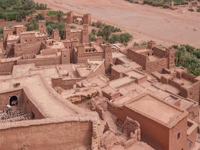 An elevated view of the earthen architecture of the Ksar of Aït Benhaddou, showing terraced rooftops, defensive towers, and a dry riverbed in the background.