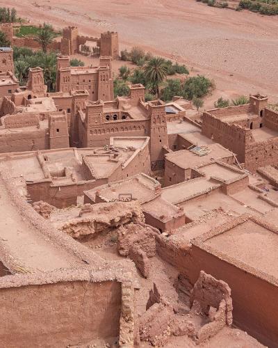 An elevated view of the earthen architecture of the Ksar of Aït Benhaddou, showing terraced rooftops, defensive towers, and a dry riverbed in the background.