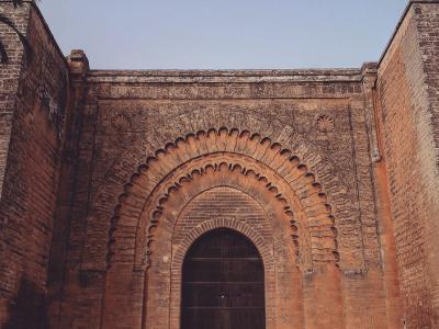 A low-angle photograph of a monumental, reddish-brown stone city gate featuring a large horseshoe arch with intricate stucco carvings and a dark wooden door, set against a clear blue sky.