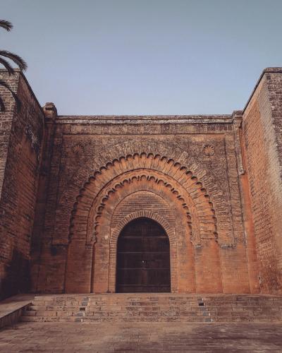 A low-angle photograph of a monumental, reddish-brown stone city gate featuring a large horseshoe arch with intricate stucco carvings and a dark wooden door, set against a clear blue sky.