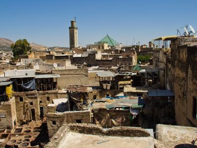 A high-angle view of a dense, historic urban district featuring traditional earthen buildings, a prominent square minaret, a green-tiled dome, and foreground rooftops with numerous satellite dishes and what appear to be dye vats.